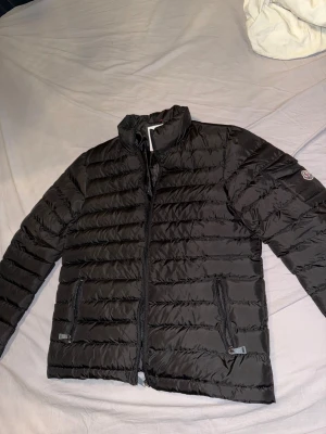 Svart Moncler pufferjacka herr - Snygg svart pufferjacka från Moncler med klassisk logga på ärmen. Jackan har ståkrage, två sidofickor med dragkedja och quiltad design. Perfekt för dig som vill ha en stilren och varm jacka med grym passform.