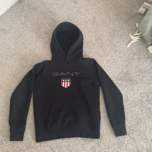 Svart hoodie från GANT med logga - Svart hoodie från GANT med stor huva och broderad logga på bröstet. Tröjan har ett stort magfack och ett snyggt emblem i rött, vitt och blått med stjärnor. Perfekt för dig som gillar klassisk streetstil och vill ha något bekvämt och stilrent. Pris kan diskuteras.