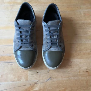 Grå/ olivgröna sneakers från lanvin - Säljer ett par grå sneakers från Lanvin med ovandel i mocka och lackad tå i mörkgrönt. Skorna har snörning och vit platt sula. Klassisk och stilren design med exklusiv känsla.  De är stora i storleken passar mig som har 43, hör av er om ni har några frågor. Bara skorna medföljer ej dust bag eller box!!