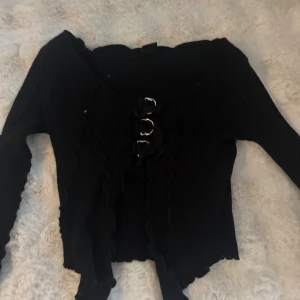 Svart croppad bolerotopp med snörning - Säljer en svart bolerotopp med långa ärmar och snygga volangdetaljer längs kanterna. Toppen är croppad och har snörning framtill med metallringar för en edgy look. Köpt på urban outfitters.