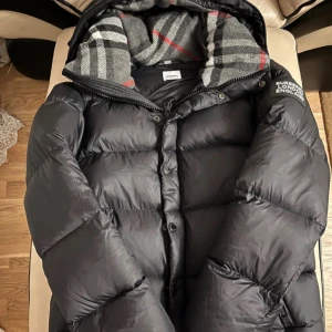 Burberry Jacka  - Svart pufferjacka från Burberry med avtagbara ärmar, så du kan bära den som väst. Jackan har huva med klassiskt rutigt Burberry-foder i grått, vitt och rött. Logotypen 'BURBERRY LONDON ENGLAND' på ärmen. Dragkedja och fickor framtill. Jackan är i storlek M men passar S också! Skicka ett meddelande vid intresse eller fler bilder! Ny utan tags. 