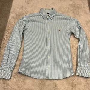 Randig blå skjorta från Ralph Lauren - Slim fit stretch oxford-skjorta från Ralph Lauren i blått och vitt randigt mönster. Klassisk button-down krage, broderad logga på bröstet och långa ärmar med manschettknappar. Skjortan är gjord i bomull med lite stretch för extra komfort.