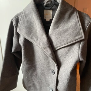 Grå kappa från Gina Tricot - Snygg grå kappa från Gina Tricot med bred krage och två stora knappar framtill. Jackan har en klassisk rak passform och är långärmad, perfekt för kyliga dagar. Materialet känns mjukt och har en ull-liknande look som ger en stilren vibe.