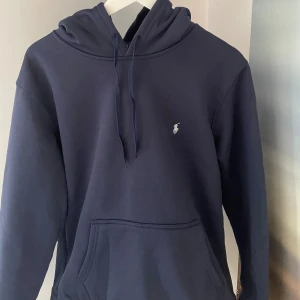Marinblå hoodie från Polo Ralph Lauren - Snygg marinblå hoodie från Polo Ralph Lauren med klassisk logga broderad på bröstet. Tröjan har huva med snörning, stor magficka och långa ärmar. Perfekt för dig som gillar stilren och enkel design med premiumkänsla.