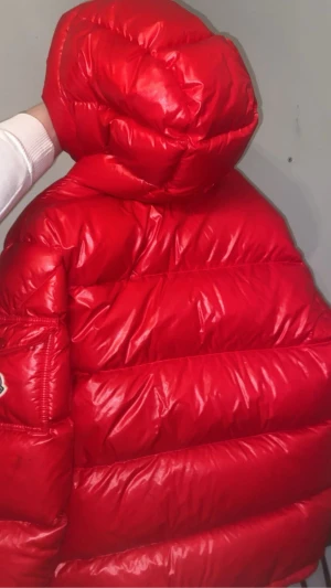 Röd Moncler encrins - Säljer en fet röd pufferjacka från Moncler med stor huva och coola detaljer som matchande röd dragkedja och knapp med Moncler-logga. Jackan har quiltad design och är tillverkad i glansig syntet med dunfyllning. Perfekt för kalla dagar och sticker ut rejält. Ny pris 19800