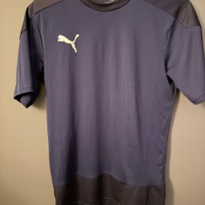 Mörkblå Puma t-shirt med logga - Snygg mörkblå t-shirt från Puma med klassisk vit logga på bröstet. T-shirten har korta ärmar och kontrastpaneler i svart vid axlar och nederkant. Materialet känns lätt och andas, perfekt för sport eller chill. Enkel och stilren design som funkar till det mesta.