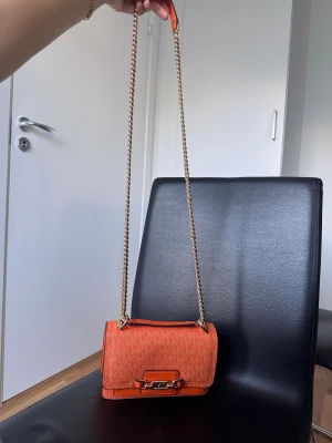 Orange axelväska från Michael Kors - Snygg orange axelväska från Michael Kors med kedjerem i guld och MK-logga framtill. Väskan har ett monogrammönster med MK-tryck över hela ytan och detaljer i skinn. Perfekt storlek för det viktigaste och ger en färgklick till din outfit.