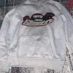 Grå Trapstar hoodie Lincoln Shooters - Säljer en grå hoodie från Trapstar med Lincoln Shooters-tryck och svart pantermotiv på bröstet. Klassisk känguruficka framtill, huva och ribbade muddar. Perfekt för dig som gillar streetwear och vill sticka ut med en unik design.