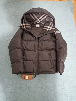 Svart Burberry pufferjacka med huva - Svart pufferjacka från Burberry med stor huva fodrad i klassiskt rutigt mönster i grått och vitt. Jackan har dragkedja och knappar framtill, samt Burberry-logga på ärmen. Snyggt quiltad design och praktiska fickor med dragkedja.
