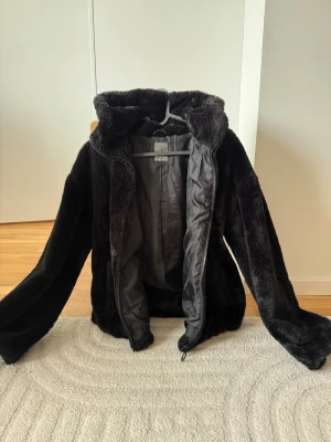 Svart teddyjacka från Primark - Mjuk och fluffig svart teddyjacka från Primark med huva och hel dragkedja framtill. Jackan har långärmade ärmar och är fodrad på insidan för extra värme. Perfekt för kalla dagar när du vill vara både chill och snygg.