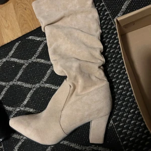 Beige Boots stövletter med klack - Snygga beige stövletter med mjukt, rynkat skaft och spetsig tå. Skorna har en stadig klack och är tillverkade i mocka för en lyxig känsla. Perfekta för dig som vill ha en trendig och stilren look. Storleken är 39-42 Dem är helt nya Aldrig använt.Finns även samma men  i färgen svart! Pris går att diskuteras!
