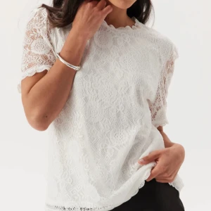 Vit spetsblus från Pieces - Supersöt vit blus från Pieces med korta ärmar och rund hals. Blusen är helt täckt av spets med blommigt mönster och har fina vågiga kanter vid ärmslut och nederkant. Aldrig använd 