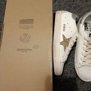 Golden Goose vita sneakers med glitterstjärna - Golden Goose sneakers i vitt skinn med beige mockadetaljer och silverfärgad häl. Sidan pryds av en glittrig guldstjärna som ger extra edge. Klassisk snörning och platt sula. Perfekta för dig som vill sticka ut med stilrena men ändå unika skor.