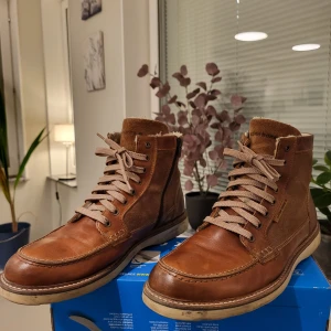 Björn Borg bruna boots i skinn och mocka - Snygga bruna boots från Björn Borg i storlek 44, tillverkade i Portugal. Skorna har en kombination av slätt skinn och mocka, beige snörning och dragkedja på sidan. Insidan är fodrad med mjukt material för extra komfort och värme. Perfekt för höst och vinter.