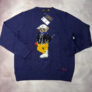Marinblå stickad tröja Polo Bear - Marinblå stickad tröja från Polo Ralph Lauren med ikoniska Polo Bear-motiv framtill. Rund hals, ribbade muddar och RL-broderi i rött vid nederkanten. Tröjan är i grovstickad bomull och har långärmad passform. Perfekt för dig som gillar klassisk streetstyle.