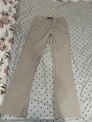 Beige bootcut kostymbyxor från BikBok - Snygga beige kostymbyxor från BikBok i storlek 34. Byxorna har bootcut-modell, hällor för bälte, fickor fram och dekorficka bak. Perfekta för en clean och stilren look. Materialet är mjukt och har en lätt stretch för extra komfort.