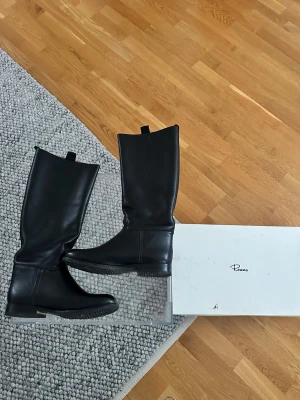 Svarta höga stövlar från Rizzo - Snygga svarta stövlar från Rizzo i äkta läder med platt sula och rund tå. Modellen är klassisk med högt skaft och diskreta dragflikar upptill. Perfekta för dig som gillar stilrena och tidlösa boots med en clean look. Mycket fint skick, inga defekter. Orginalpris: 2795kr