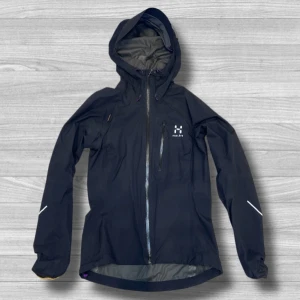 Svart Haglöfs vindjacka XS dam - Svart vindjacka från Haglöfs för dam i storlek XS. Jackan har huva, långa ärmar och reflekterande detaljer på ärmarna. Tillverkad i polyamid med Gore-Tex-membran, perfekt för blåsiga och regniga dagar. Dragkedja framtill och diskret logga på bröstet.