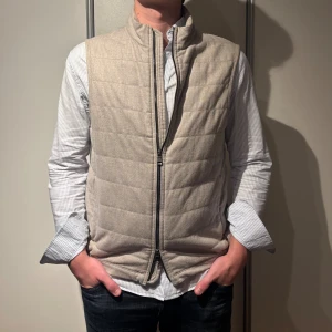 Beige linneväst Oscar Jacobson - Snygg beige linneväst från Oscar Jacobson med quiltad design och hög krage. Västen har två sidofickor med dragkedja och stilren look som funkar till skjorta eller hoodie. Perfekt för lager-på-lager under höst och vinter. Dragkedja framtill för enkel på- och avtagning.