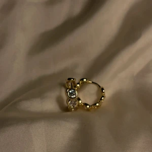 Guldiga hoops med stenar - Säljer ett par snygga guldiga hoops med glittrande stenar runt hela örhänget. De är runda och har en cool bubblig design som verkligen sticker ut. Perfekta för dig som vill ha något extra till din look.