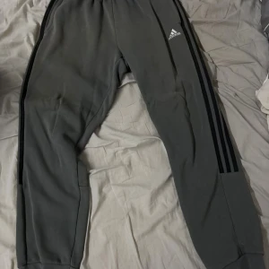 Grå Adidas mjukisbyxor M - Grå mjukisbyxor från Adidas med klassiska svarta ränder längs sidorna och Adidas-logga på låret. Byxorna har elastisk midja och är gjorda i mjukt bomullsmaterial, perfekta för chill eller träning. Snygg tapered passform som sitter skönt.