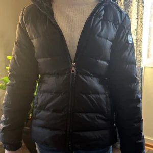 Svart pufferjacka från Authentic Athletic - Svart pufferjacka från Authentic Athletic Apparel med snyggt emblem på ärmen. Jackan har dragkedja framtill, huva med snörning och quiltad design. Perfekt för kalla dagar och har en modern, loose passform som funkar till jeans eller hoodie.