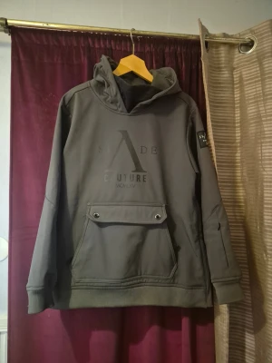 Olivgrön/ grå skidjacka från Slade Couture - Snygg olivgrön (men ser nästan frå ut) skidjacka från Slade Couture med stor logga framtill och patch på ärmen. Tröjan har huva, lång ärm och en stor ficka med knappdetaljer. Perfekt för dig som åker skidor då den står emot blåst och har praktiska fickor. Den är väldigt bekväm så den passar även som streetwear 