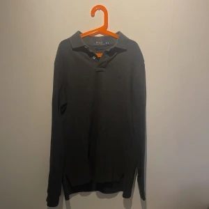 Mörkgrå pikétröja från Polo Ralph Lauren - Säljer en mörkgrå långärmad pikétröja från Polo Ralph Lauren. Tröjan har klassisk krage, knäppning vid halsen och den ikoniska lila broderade loggan på bröstet. Tillverkad i mjuk bomull som är skön mot huden.