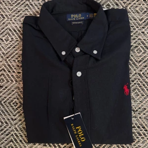 Svart skjorta från Polo Ralph Lauren - Svart skjorta från Polo Ralph Lauren med klassisk krage och knappar framtill. Slim fit-modell med rött broderat Polo-logo på bröstet. Skjortan är i mjuk bomull och har en stilren look som funkar till många olika outfits.