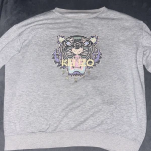 Grå Kenzo sweatshirt med tigertryck - Säljer en grå sweatshirt från Kenzo med ikoniskt broderat tigerhuvud i pastellfärger och Kenzo Paris-text på bröstet. Tröjan har rund hals, långärmade och är tillverkad i mjuk bomull. Perfekt för dig som gillar streetstyle och vill sticka ut med ett coolt motiv.