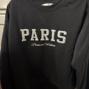 Svart sweatshirt med Paris-tryck - Svart sweatshirt med stort Paris-tryck i vitt på bröstet. Rund hals och långa ärmar. Mjuk bomullskänsla och relaxed fit, perfekt för chill dagar