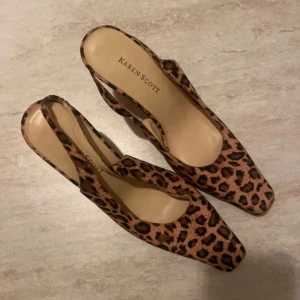 Leopardmönstrade pumps från Karen Scott - Säljer ett par snygga pumps från Karen Scott med leopardmönster i bruna och svarta toner. Skorna har spetsig tå, öppen häl med rem och klack. Materialet är skinnliknande och de har en elegant form som sticker ut.