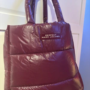 Vinröd puffad axelväska Marc Jacobs - Snygg vinröd puffad axelväska från Marc Jacobs med quiltad look och glansig finish. Väskan har breda, vadderade handtag och tydlig logga framtill. Perfekt för dig som vill sticka ut med en trendig och rymlig väska. Väskan är sprillans ny och aldrig använd. 