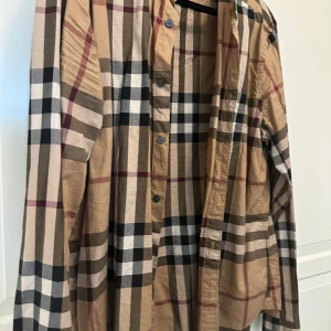 Burberry Brit rutig skjorta XL - Snygg rutig skjorta från Burberry Brit i klassiska beige, svarta och röda toner. Skjortan har långa ärmar, knappar framtill och är gjord i mjuk bomull. Perfekt för dig som gillar ikoniska mönster och vill sticka ut med en tidlös look.