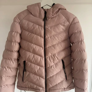 Ljusrosa pufferjacka från FB Sister - Säljer en ljusrosa pufferjacka från FB Sister i storlek S. Jackan har huva, svarta dragkedjor och två fickor framtill. Insidan är fodrad med svart fleece för extra värme. Perfekt för kalla dagar och har en snygg, quiltad look.