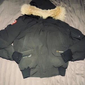 Svart dunjacka med päls från Canada Goose - Svart dunjacka från Canada Goose med huva och fluffig ljus päls runt kragen. Jackan har ribbade muddar, dragkedja framtill och flera fickor. Klassisk Canada Goose-märke på ärmen och snygga detaljer. Perfekt för kalla vinterdagar.