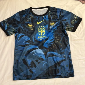 Blå Brasil Nike fotbollströja leopard - Säljer en unik Brasilien fotbollströja från Nike med ett stort blått leopardmotiv över hela tröjan. Trycket har gula detaljer och Brasil-logga på bröstet. Tröjan är kortärmad med svart krage och är tillverkad i lätt, ventilerande syntetmaterial.