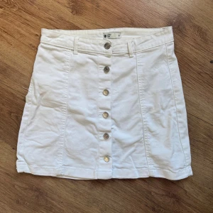 Vit kortkjol med knappar från Gina Tricot - Supersnygg vit kortkjol från Gina Tricot med raka linjer och silverfärgade knappar framtill. Kjolen har hällor för bälte. 