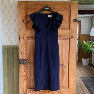 Marinblå långklänning från Coast - Snygg marinblå långklänning från Coast med v-ringad hals och volangdetaljer vid axlarna. Klänningen har en elegant, figurnära passform och är ärmlös. Perfekt för dig som vill ha en stilren look med lite extra edge.