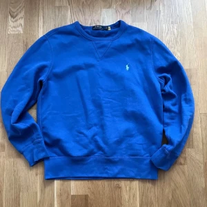Blå sweatshirt från Polo Ralph Lauren - Klassisk blå sweatshirt från Polo Ralph Lauren med rund halsringning och broderad logga på bröstet. Tröjan har långa ärmar och ribbade muddar vid ärmslut och nederkant. Perfekt för en chill och stilren look.