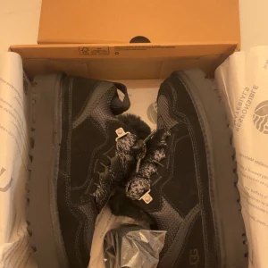  UGG Lowmel - Säljer ett par svarta UGG boots med grov sula och pälsfoder. Skorna har snörning med mönstrade svarta och gråa skosnören, robust yttersula och detaljer i mocka och syntet. Perfekta för kalla dagar och har en cool, chunky look. 