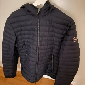 Mörkblå Colmar dunjacka - Mörkblå vadderad jacka med dragkedja fram och luva. Storlek 50 skulle att den passar som M/L men det finns tabeller om man söker pris går att diskutera. Jackan är använd en vinter och har ett litet hål vid axeln därav priset. Tveka inte på att skriva :)
