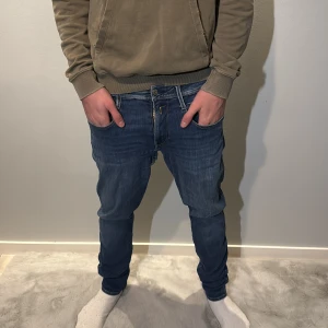 Replay Anbass jeans blå tapered - Säljer ett par snygga Replay Anbass jeans i blå tvätt med klassiska fem fickor och Replay-logga vid fickan. Jeansen har tapered passform, normal midja och är tillverkade i mjukt jeanstyg med snygga slitningar. Perfekt för dig som gillar stilrena och bekväma jeans. Modellen på bilden är 190cm lång och väger 75kg