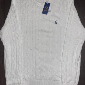 Vit kabelstickad tröja Polo Ralph Lauren - Kabelstickad vit tröja från Polo Ralph Lauren med klassisk rund halsringning och broderad marinblå logga på bröstet. Tröjan har ribbade muddar och är tillverkad i mjuk bomull, perfekt för en clean och stilren look.