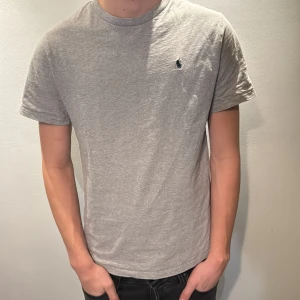 Grå t-shirt från Ralph Lauren - Snygg grå t-shirt från Ralph Lauren med klassisk broderad logga på bröstet. T-shirten är i mjuk bomull och har en normal passform med korta ärmar och rund hals. Perfekt till jeans eller shorts för en clean och enkel stil. Modellen är 186 cm och väger 70 kg 