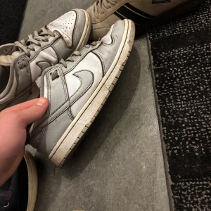 Nike Dunk Low sneakers vit/grå - Nike Dunk Low sneakers i vitt skinn med klassisk rund tå och snörning. Snygg retrostil med låg profil och ikonisk swoosh på sidan. Perfekta för dig som gillar streetwear och vill ha en clean look.