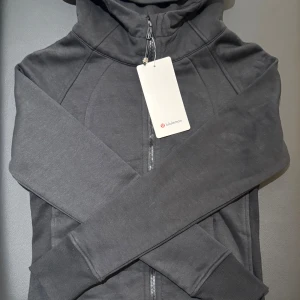 Svart hoodiejacka från Lululemon i storlek 6 - Svart hoodiejacka från Lululemon med dragkedja framtill och broderad logga på huvan. Hoodien är tillverkad i mjuk bomull och polyester, har långa ärmar och ribbade muddar. Perfekt för chill dagar eller när du är på språng.