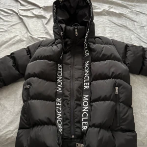 Svart dunjacka från Moncler med huva - Säljer en svart dunjacka från Moncler med stor huva och tydliga logotexter längs dragkedjan. Jackan har två fickor med dragkedja och klassisk Moncler-logga på ärmen. Perfekt för kalla dagar och riktigt snygg streetstil.