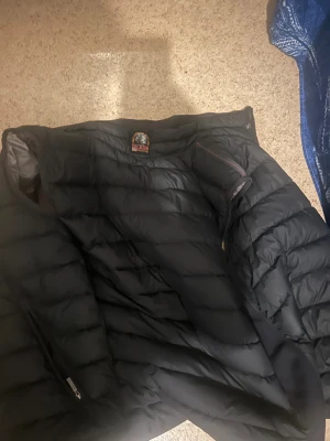 Svart dunväst från Parajumpers - Snygg svart dunväst från Parajumpers i storlek S. Västen har quiltad design, hög krage och dragkedja framtill samt två sidofickor med dragkedja. Insidan är grå och har coola detaljer som gul rem vid halsen. Perfekt för lager-på-lager och streetstyle.