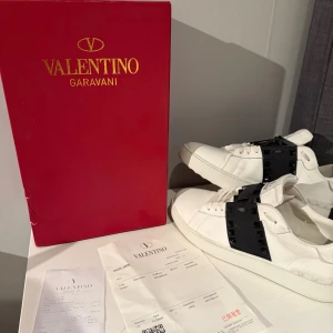 Valentino Garavani Rockstud sneakers - Säljer ett par vita Valentino Garavani Rockstud sneakers med svart band och ikoniska pyramidnitar. Skorna har rund tå, platt sula och snörning. Tillverkade i skinn med coola detaljer som ger en edgy vibe. Box och kvitto medföljer.  